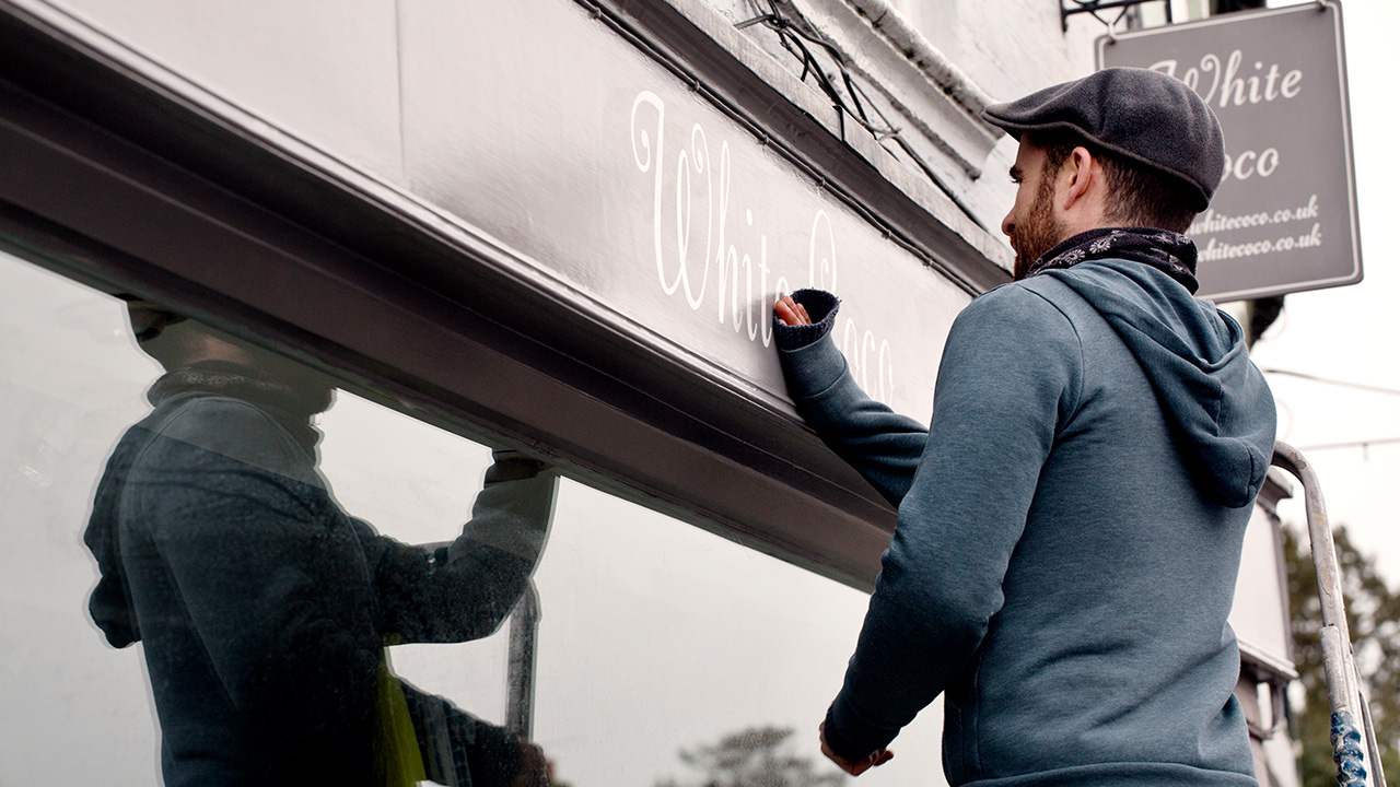 A man on a ladder fixing a painted name sign onto a bracket on a shopfront
